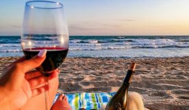 Hand holding a glass of wine with a bottle of wine on the beach.
