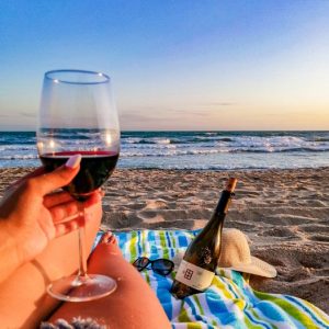 Hand holding a glass of wine with a bottle of wine on the beach.